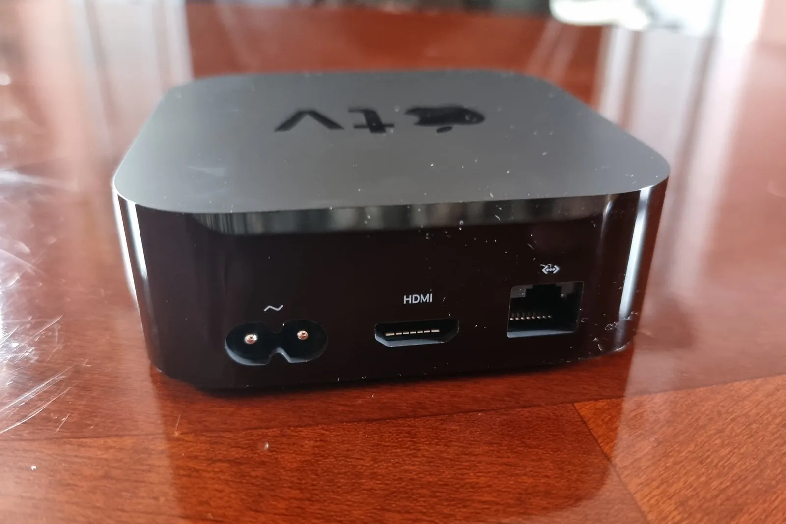 The back panel of a black Apple TV box on a polished wooden table: a figure-eight power socket, an HDMI port and an Ethernet port marked with a network symbol.