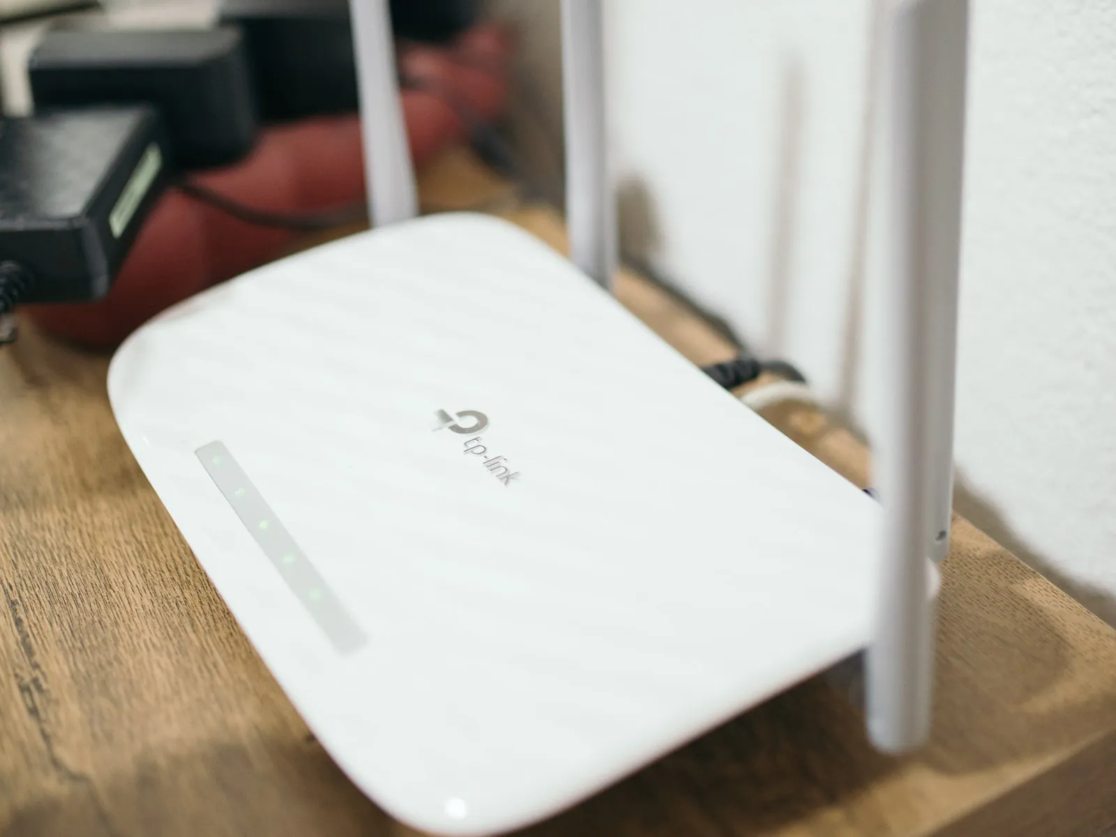 A white router with four antennas and the TP-Link logo on its lid, status lights lit, on a wooden side table, with power adapters on a red cloth behind it.