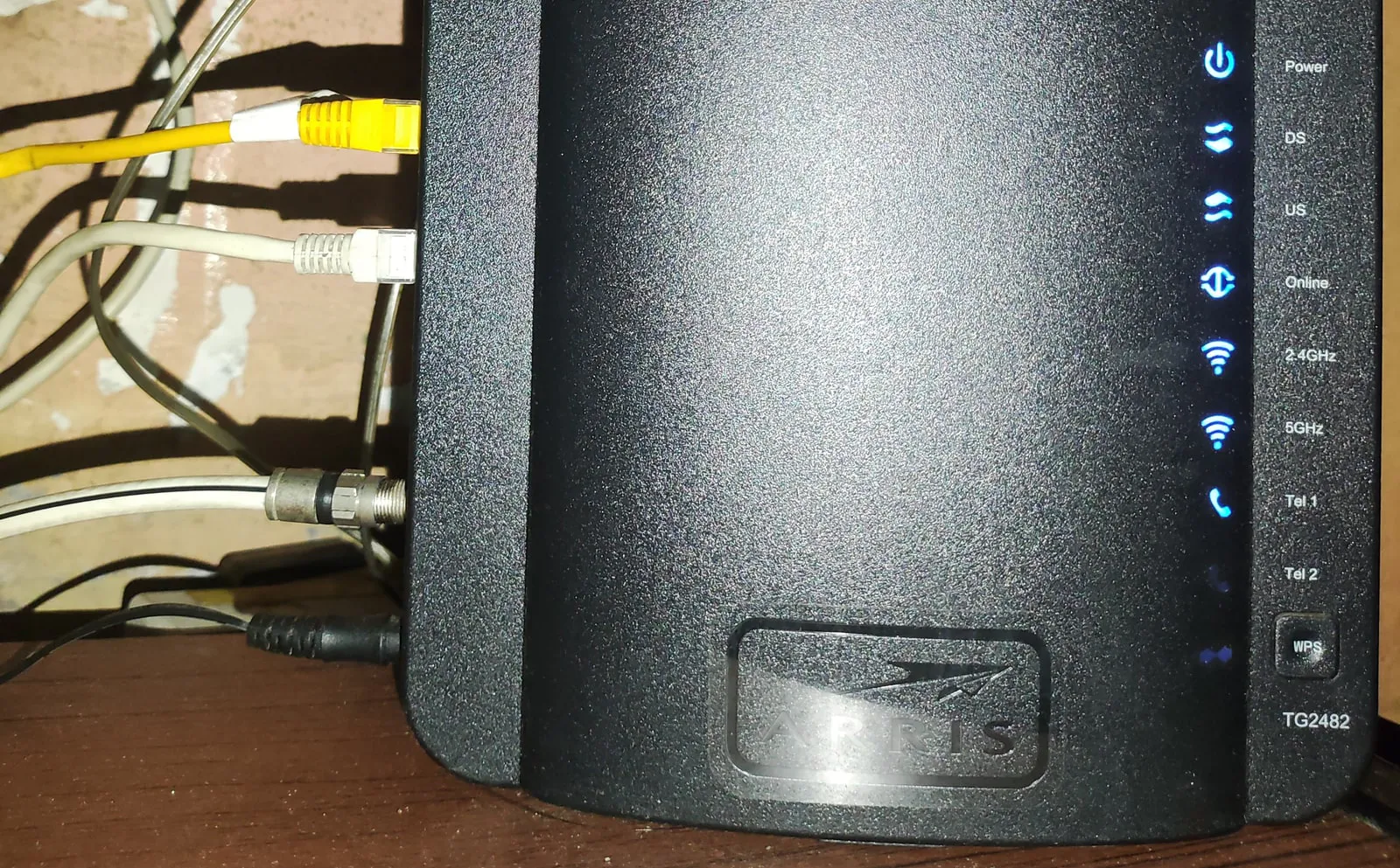 The front of a black cable gateway with a column of blue status lights labelled Power, DS, US, Online, 2.4GHz, 5GHz and Tel 1, with a coax cable and two Ethernet cables plugged into its side.