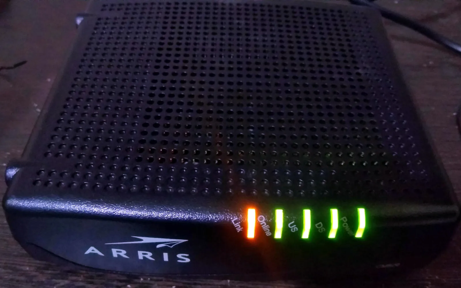 A black ARRIS cable modem on a wooden surface, its front status lights lit: Link in amber, then Online, US, DS and Power in green.