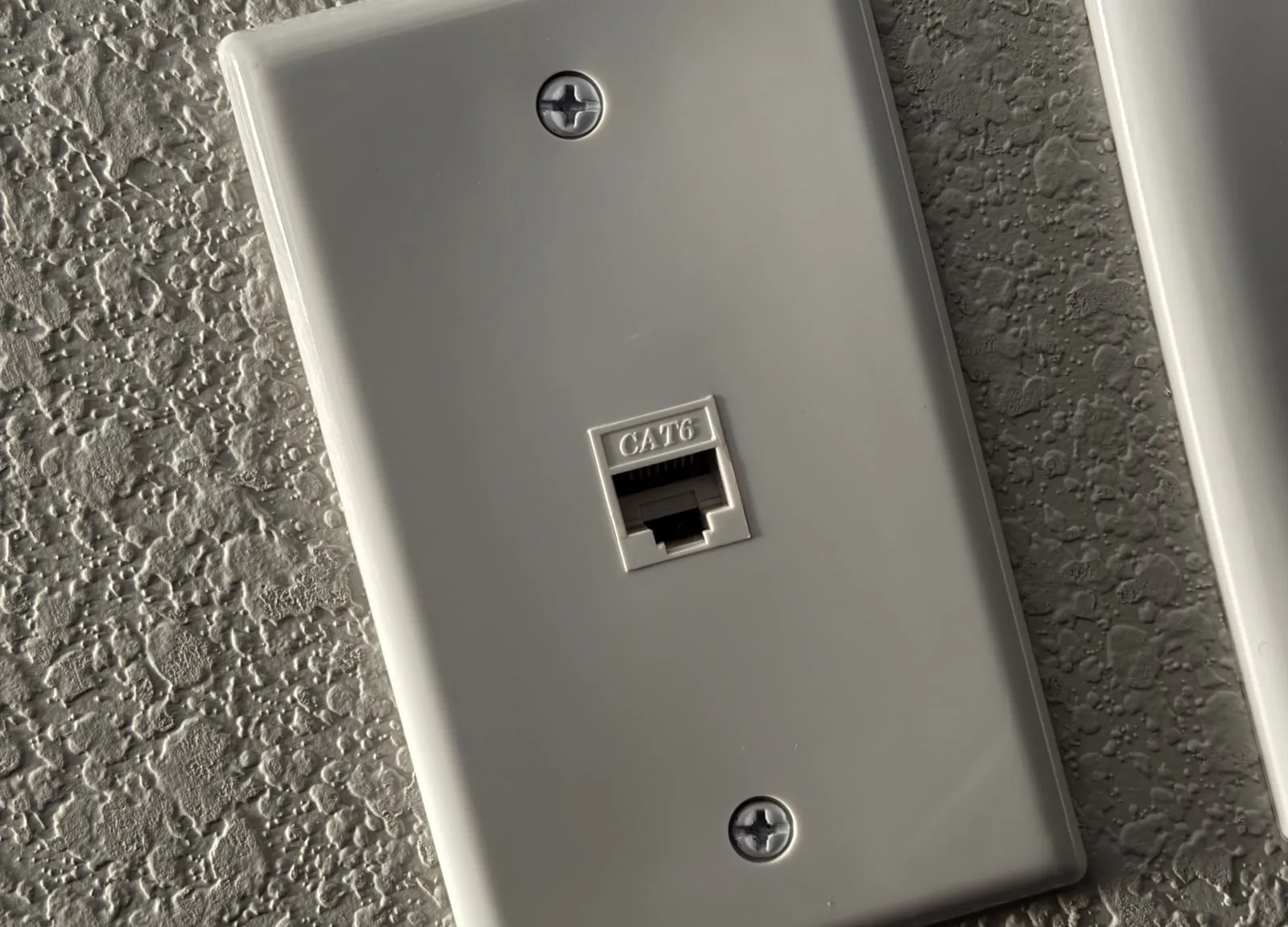 A white single-port wall plate on a textured wall; the Ethernet jack in the middle is marked CAT6.