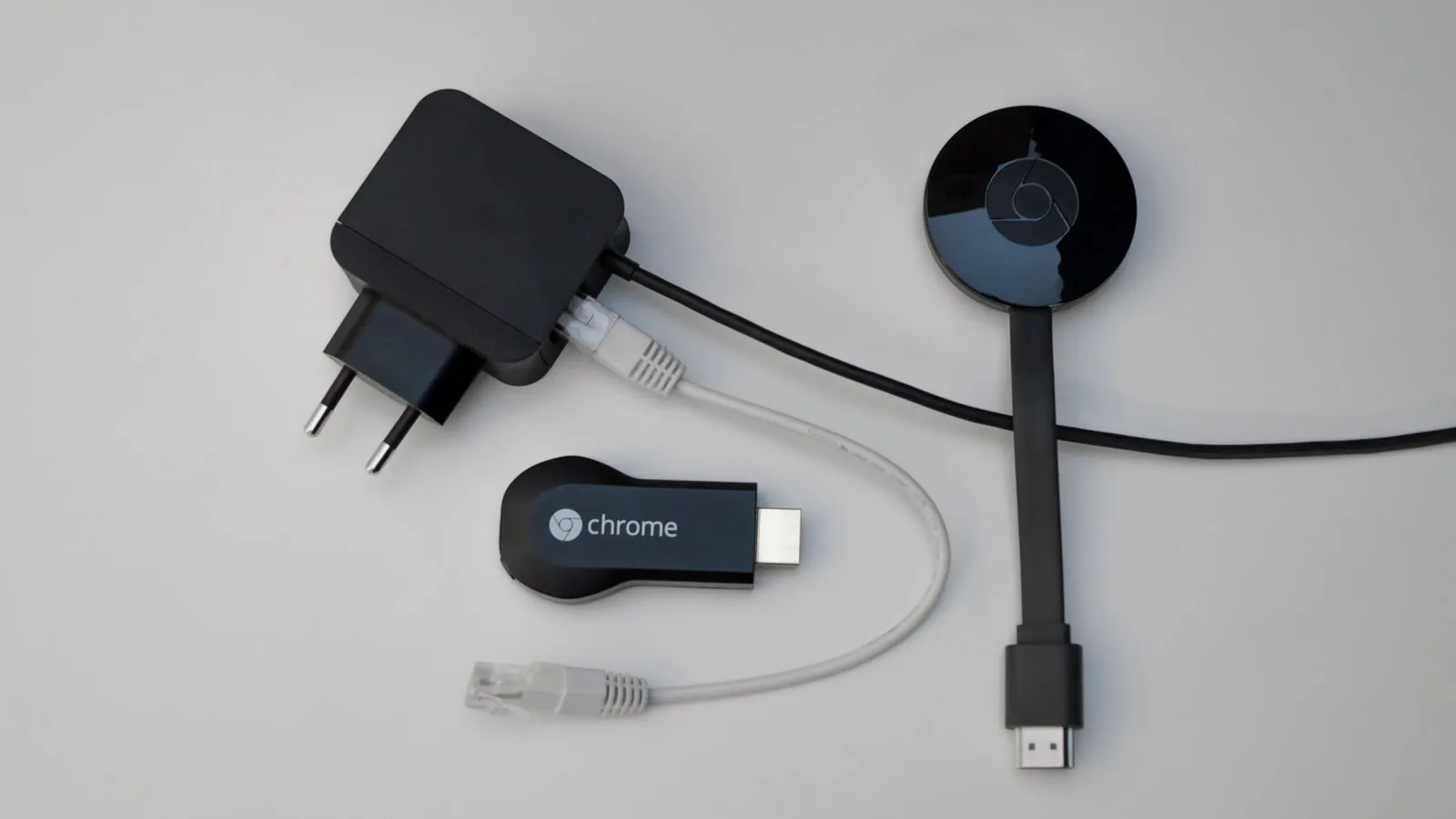 A black Chromecast power adapter with a grey Ethernet cable plugged into it, beside two Google Chromecast streaming devices on a white table.