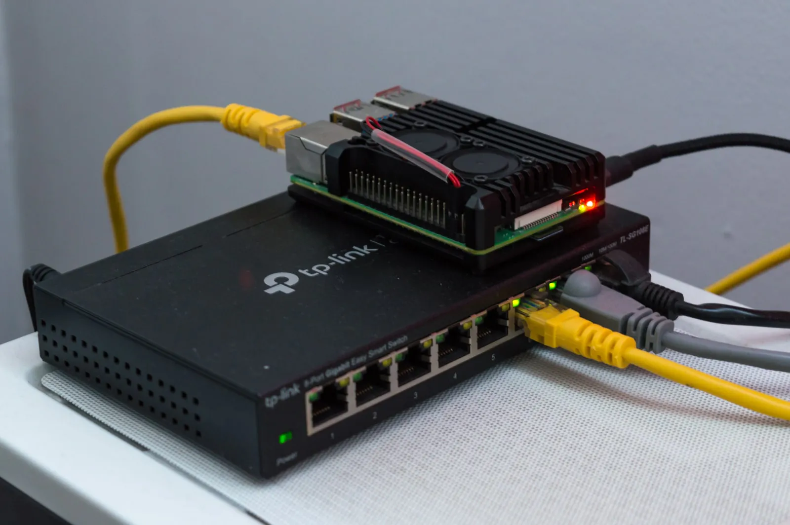 A black eight-port desktop switch with yellow, grey and black Ethernet cables plugged into its right-hand ports and green link lights, with a Raspberry Pi in a black fan case resting on top.
