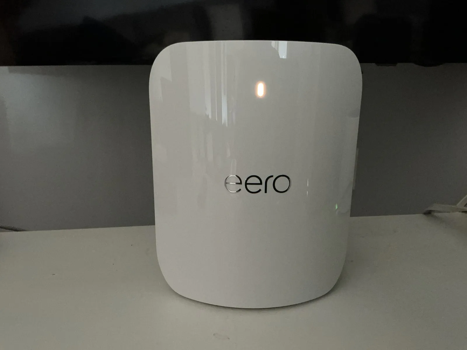 A white eero mesh unit with a glossy curved front and a lit status light, standing on a white shelf in front of a television.