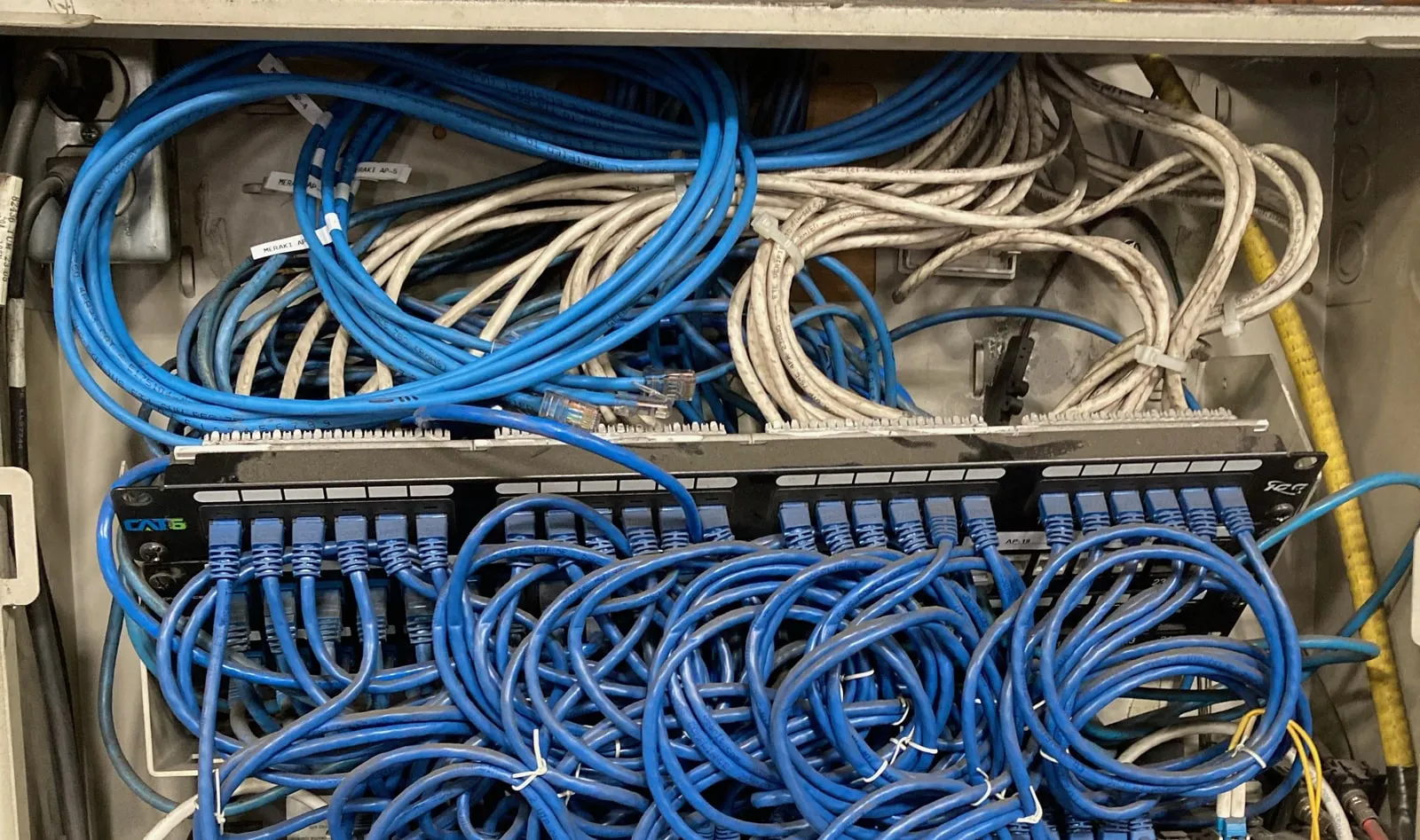Inside a wall-mounted network enclosure, dozens of blue and grey Ethernet cables, some tagged 'Meraki AP', run into the back of a long black Cat6 patch panel, with a tangle of short blue patch cords plugged into its front.