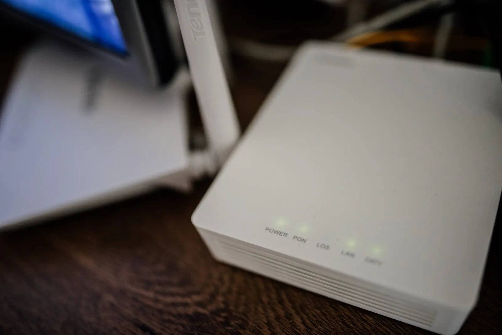 A small white fiber terminal on a wooden desk with green lights labelled Power, PON, LOS, LAN and CATV, and a white Wi-Fi router with an antenna out of focus behind it.
