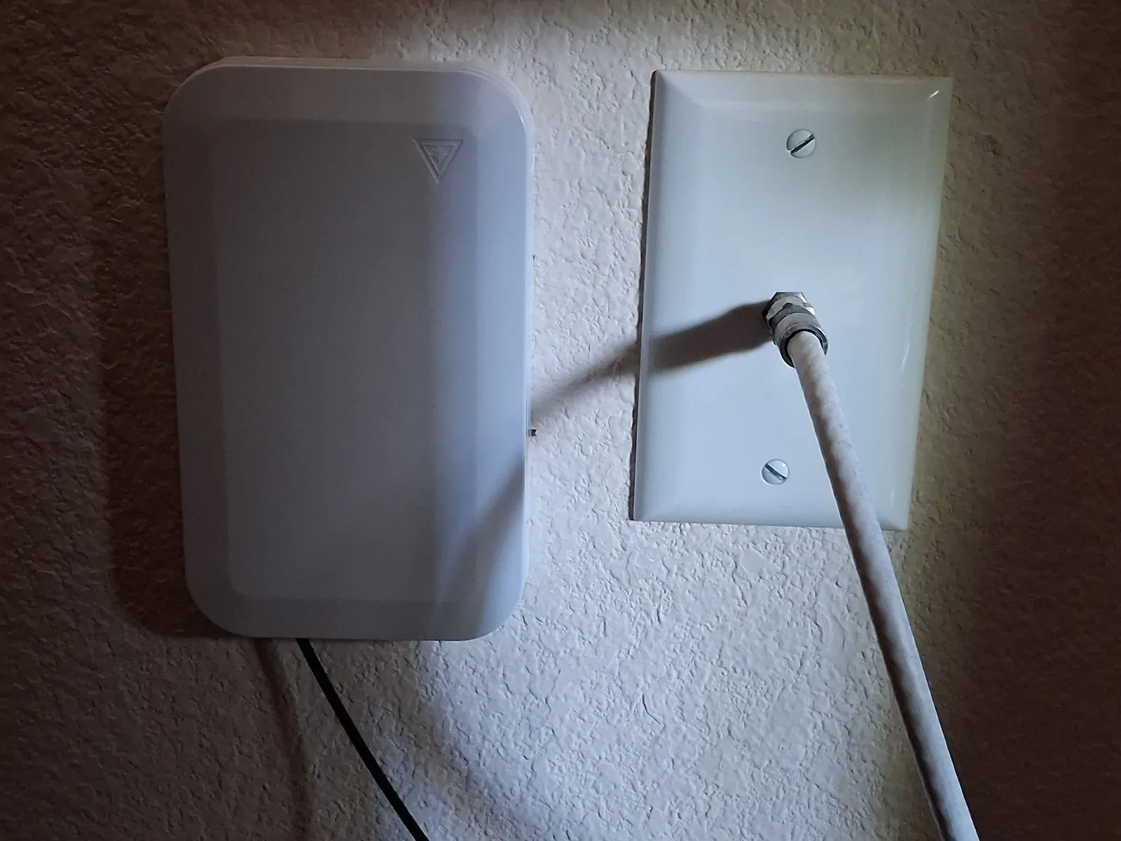 Two white fittings side by side on a textured wall: on the left a fiber provider's wall box with a thin black cable leaving its base, on the right a wall plate with a coaxial cable screwed into it.