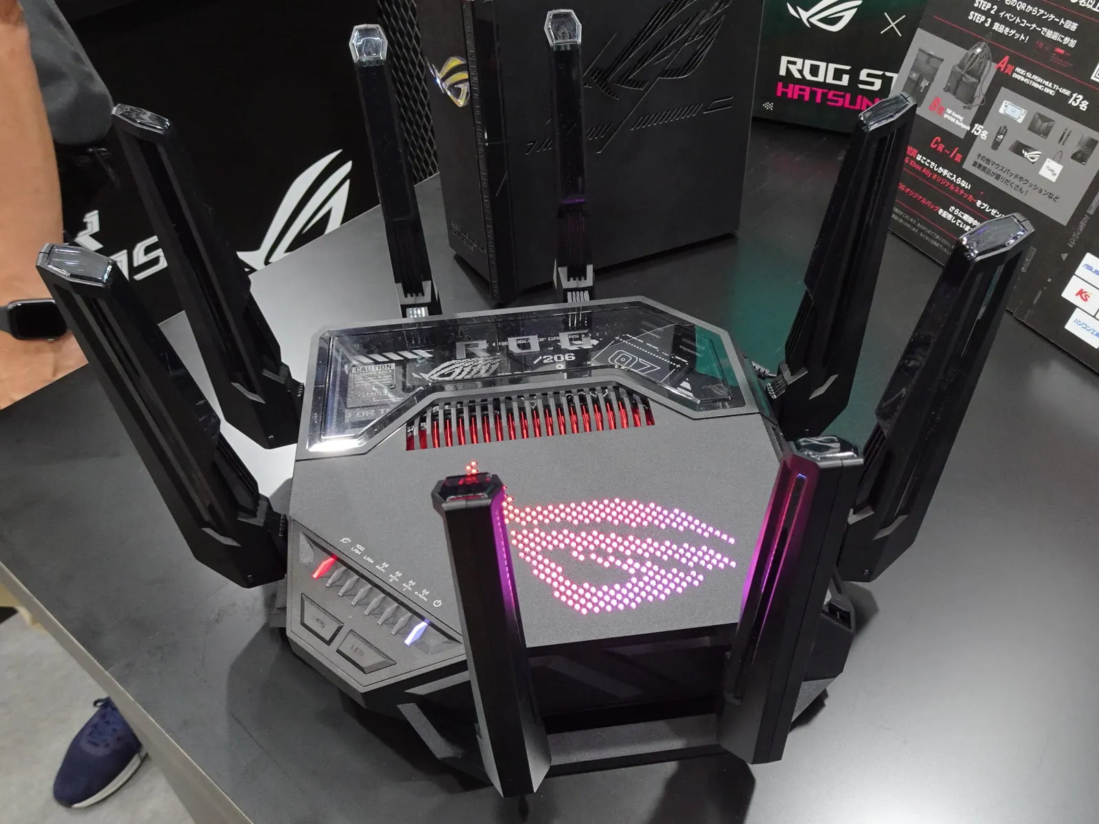A black ASUS ROG gaming router with eight angular antennas, a glowing pink ROG logo made of LEDs and red-lit vents, on a demo table at a game show.