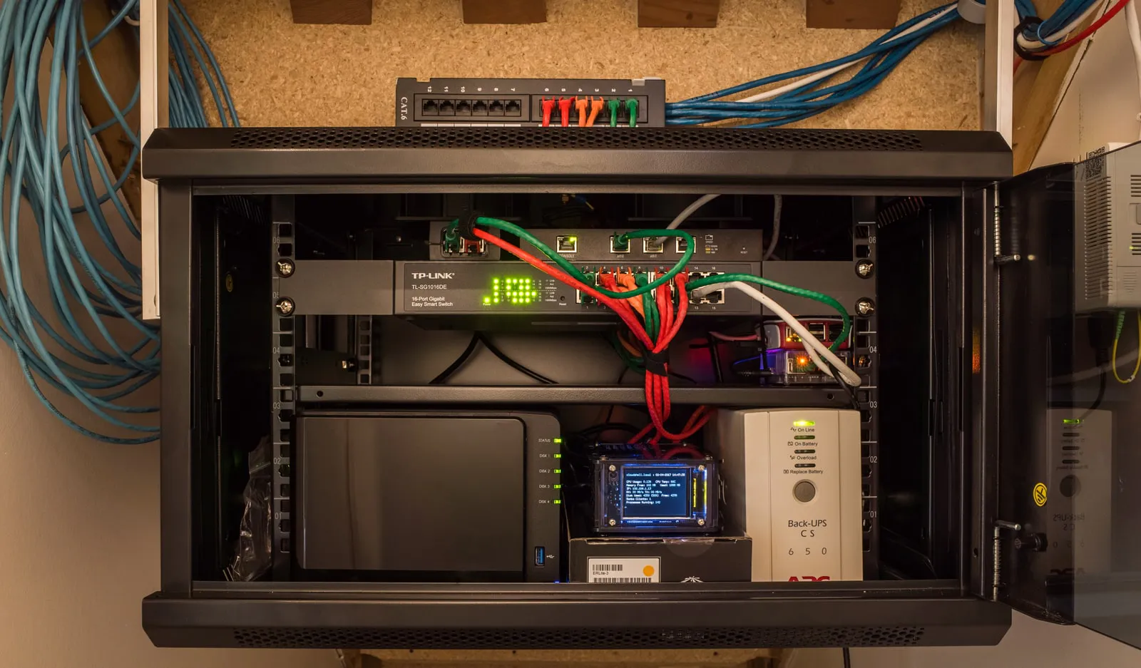A small wall-mounted network cabinet with its door open: a patch panel above it, a 16-port switch with red, green and white patch cables, a four-bay NAS at the bottom left, a small status screen and a UPS at the bottom right.