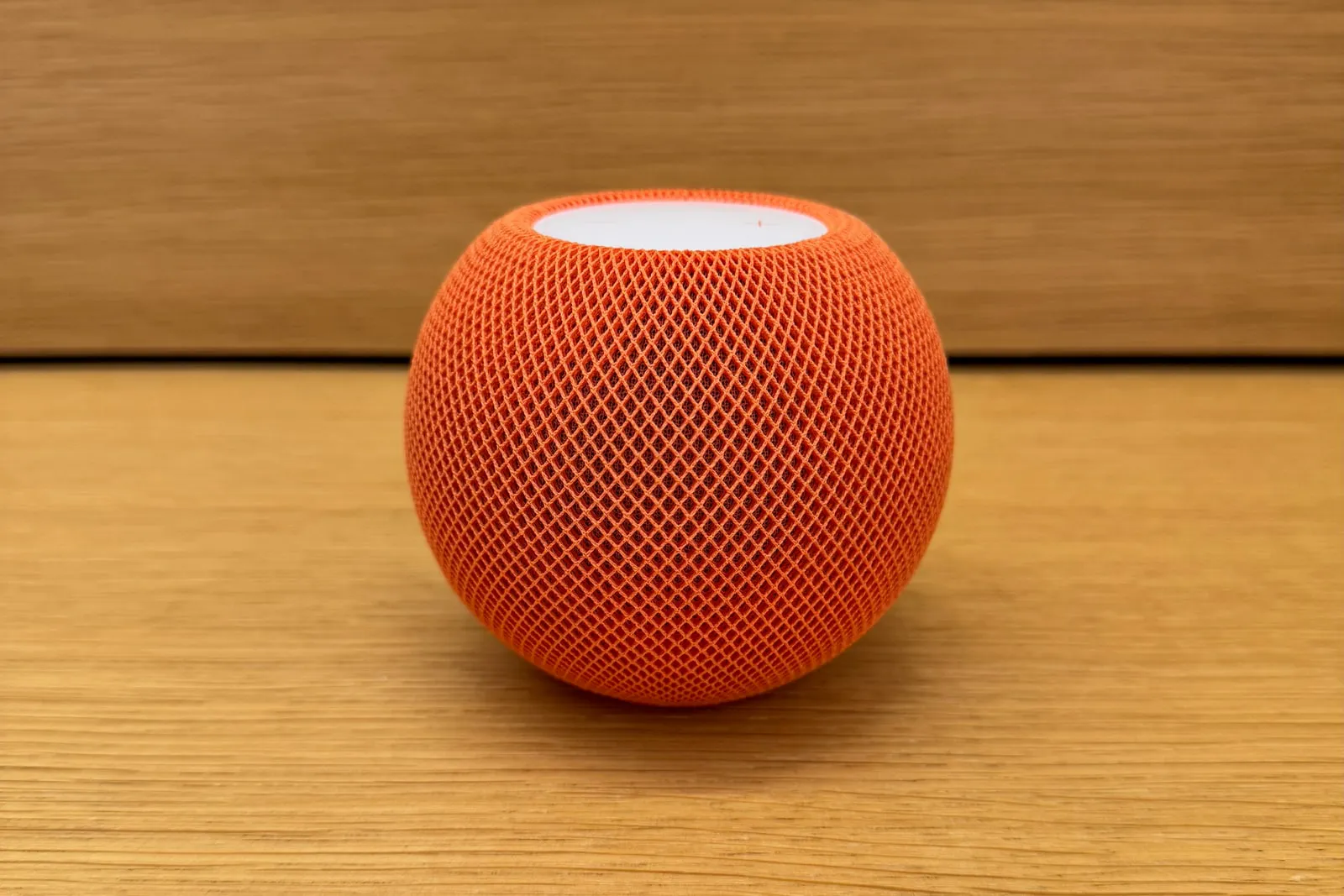 An orange HomePod mini, a small round speaker covered in mesh with a white top, standing on a light wooden surface.