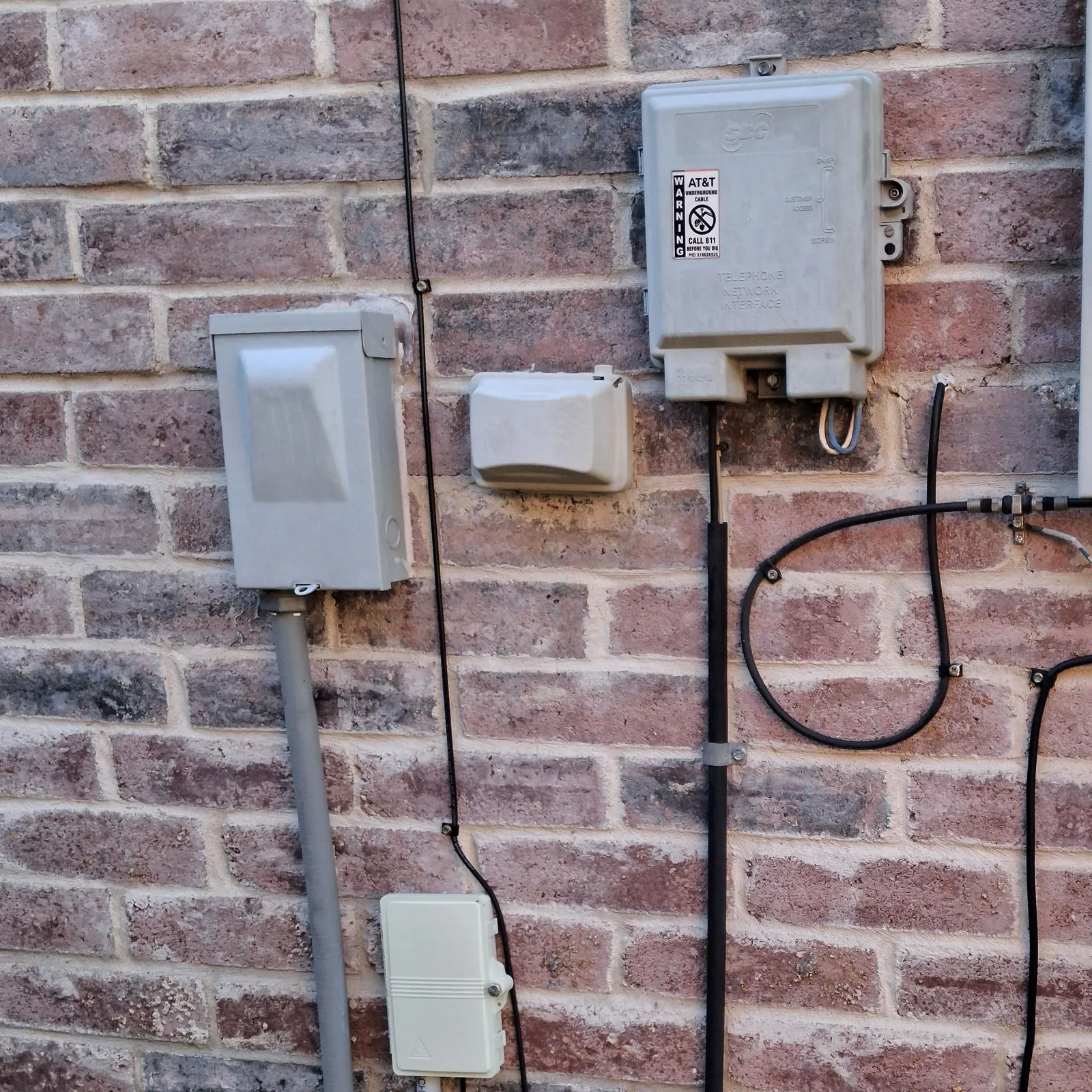 The outside brick wall of a house with a grey box labelled Telephone Network Interface, a smaller white box on a thin black cable, black coaxial lines with a connector, and an electrical box with conduit.
