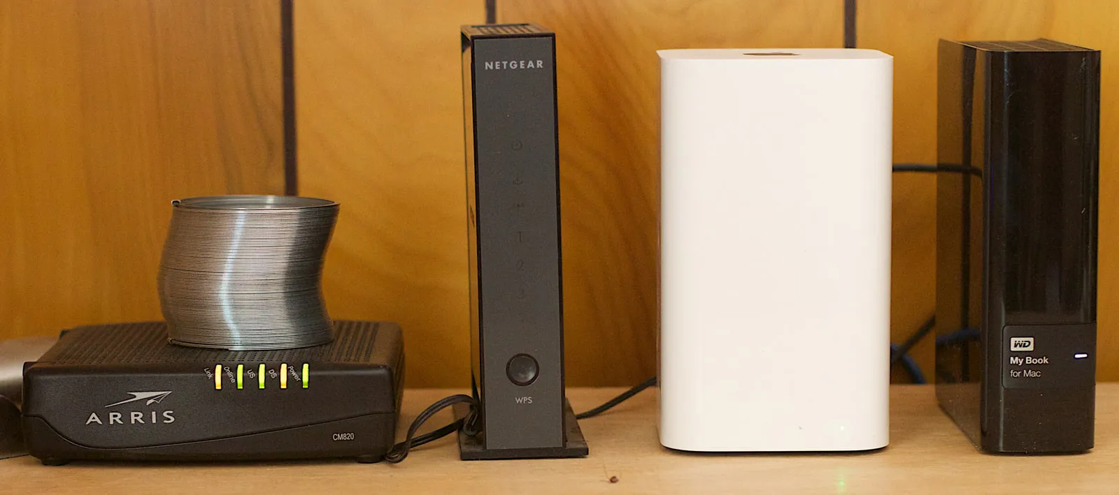 A wooden shelf with a black cable modem (a metal Slinky resting on it), an upright black Netgear router, a white Apple AirPort Extreme and a black Western Digital external drive.