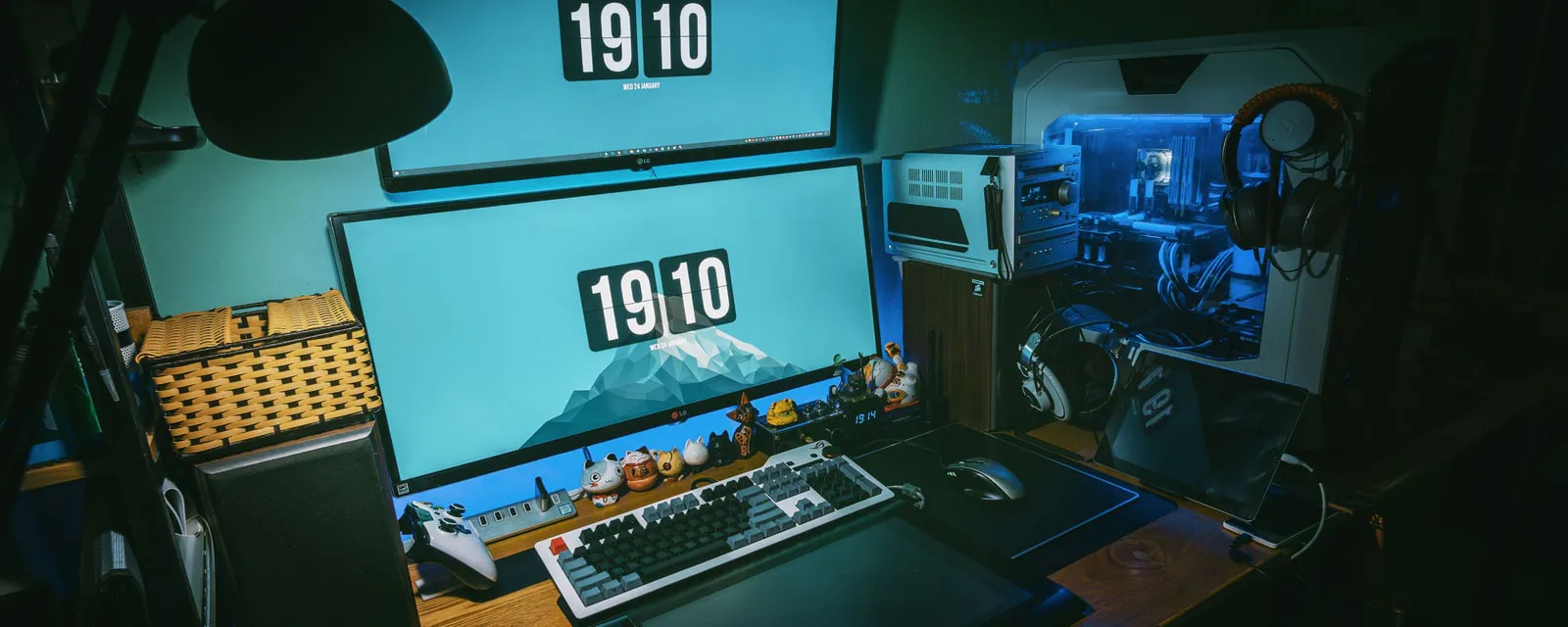 A dimly lit home desk with two monitors, a mechanical keyboard, a game controller and a desktop PC with a lit glass side panel, next to a small stereo and headphones.