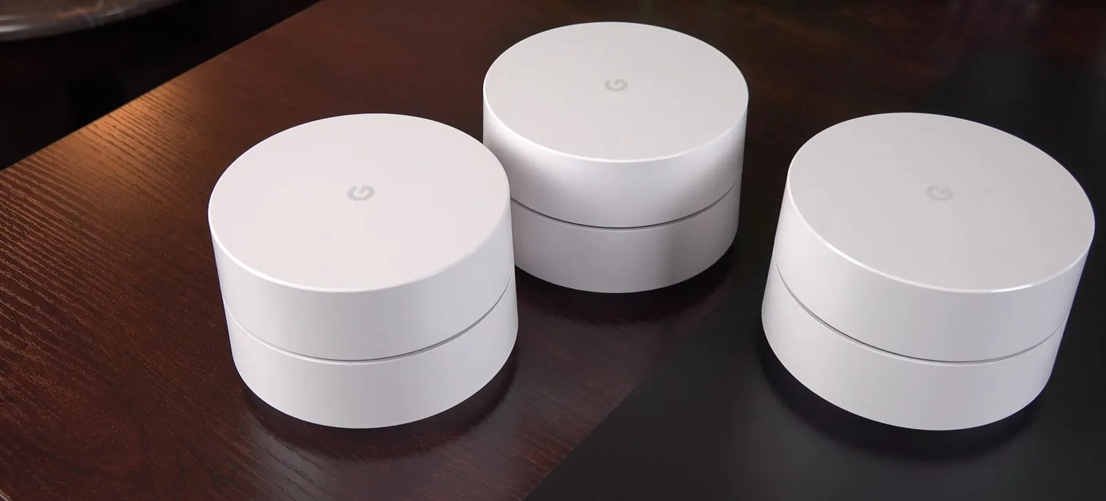 Three round white Google Wifi units side by side on a dark wooden table.