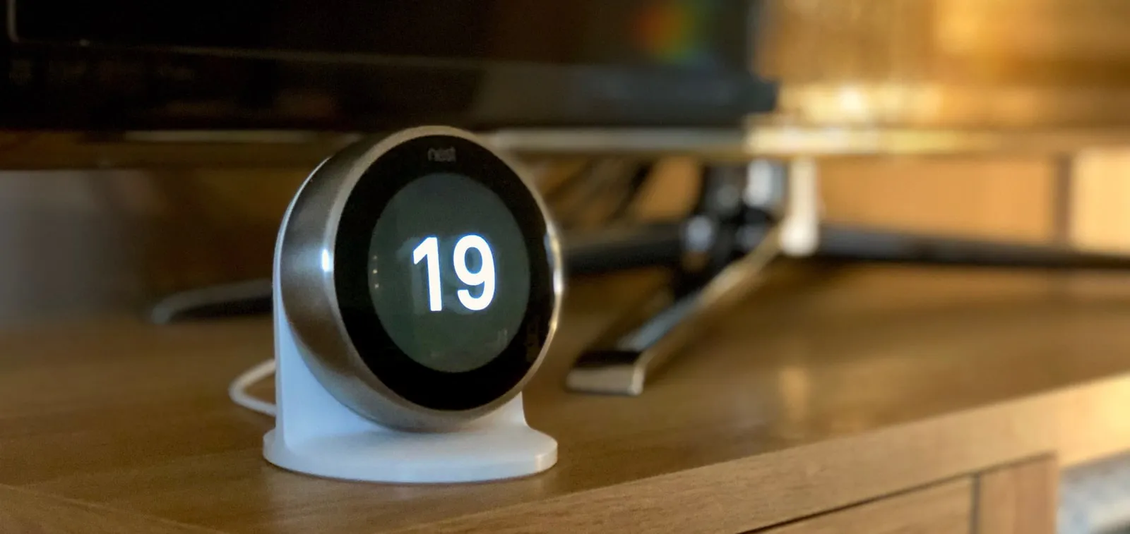 A round Nest thermostat showing 19 on its display, on a white table stand on a wooden TV cabinet, with the foot of a TV behind it.