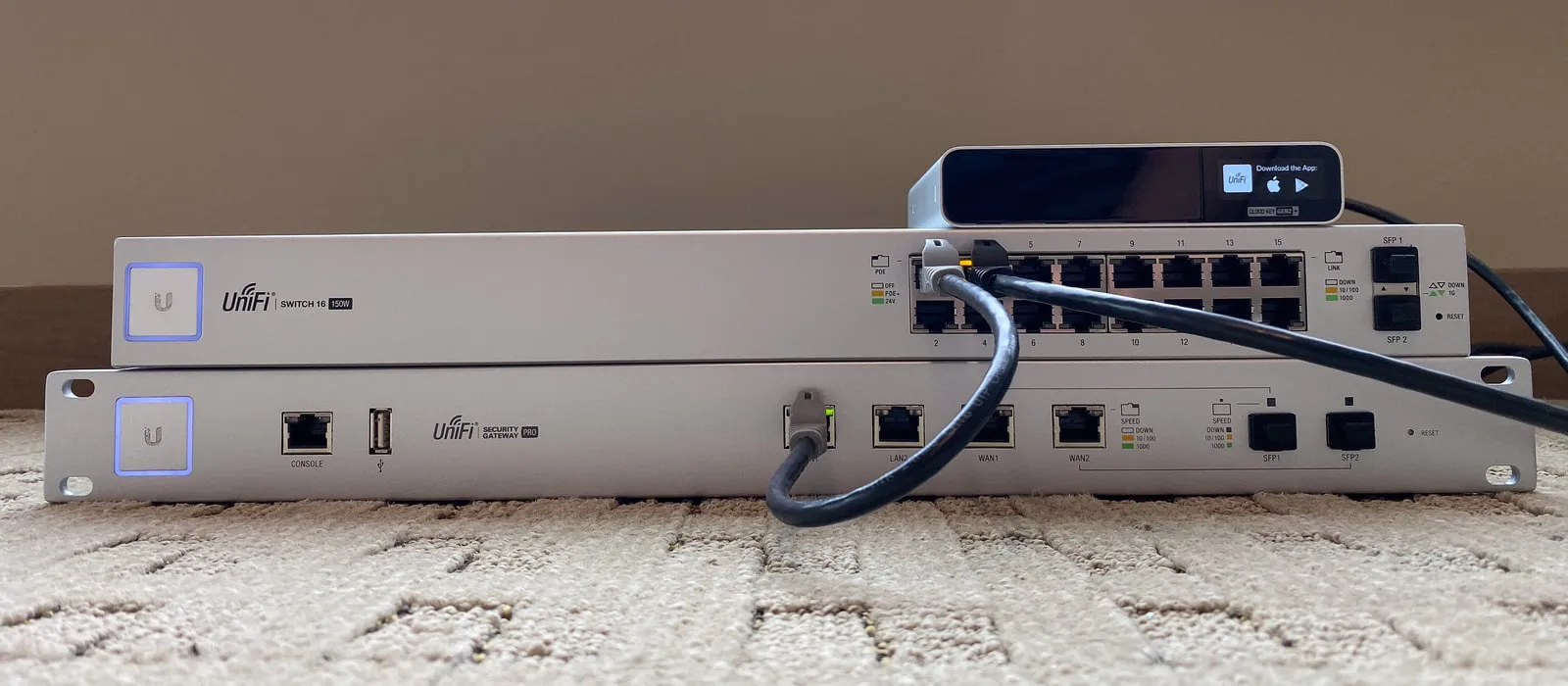 Two white rack-mount Ubiquiti UniFi units stacked on a carpet: a security gateway at the bottom, a 16-port switch with patch cables above it, and a small UniFi device with a status screen on top.