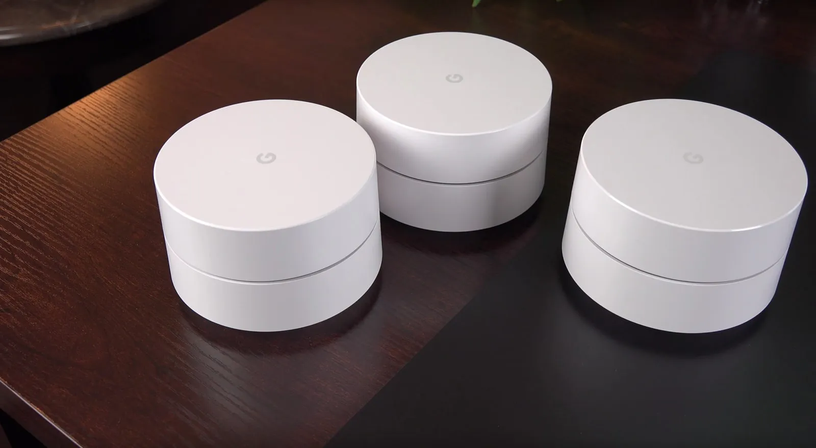 Three round white Google Wifi mesh points standing on a dark wooden table.