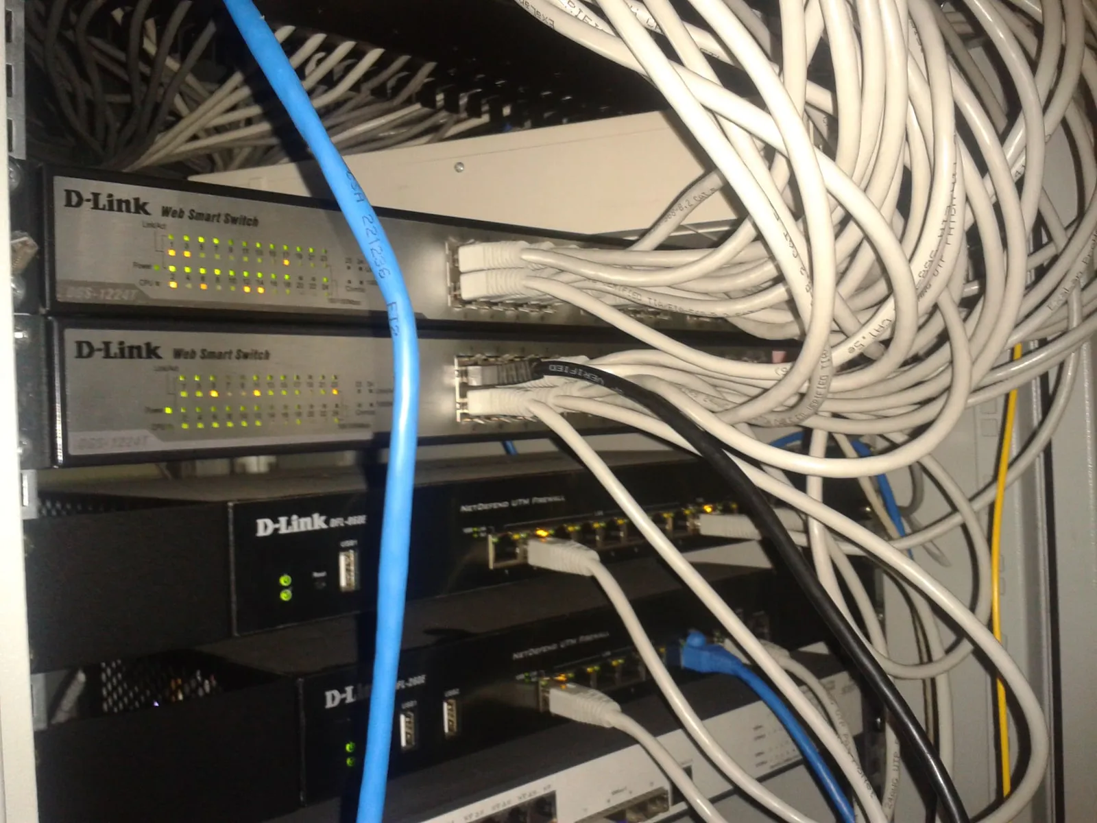 A network rack with two grey D-Link Web Smart switches, their port lights lit and grey patch cords plugged in, above two black D-Link NetDefend firewall appliances.