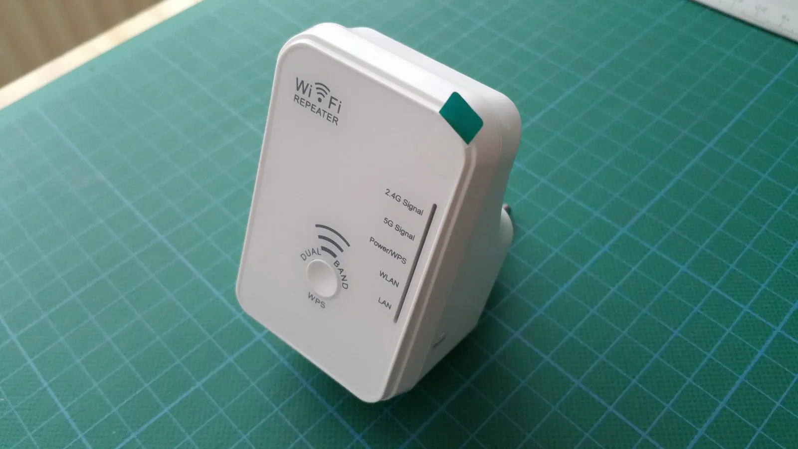 A small white plug-in Wi-Fi repeater labelled WiFi Repeater, with signal lights for 2.4 and 5 GHz, a WPS button and a mains plug on its back, standing on a green cutting mat.