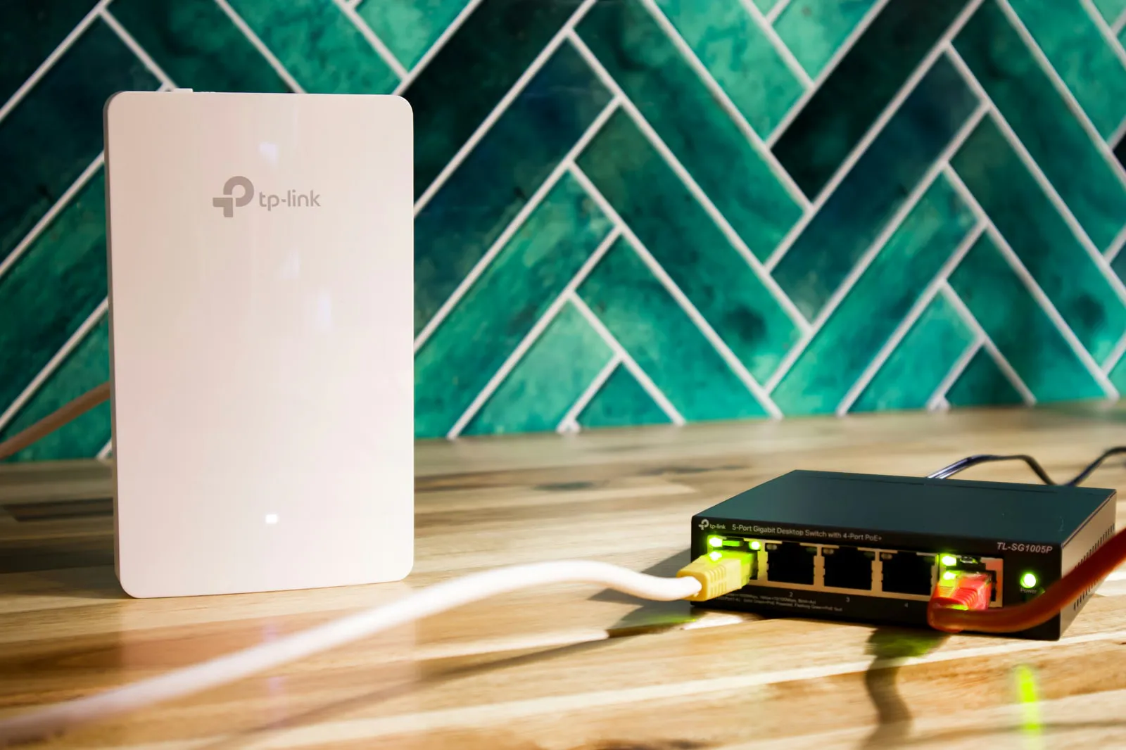 A five-port desktop network switch with green port lights and yellow and red cables plugged in, next to a flat white wall-mount Wi-Fi access point on a wooden worktop.