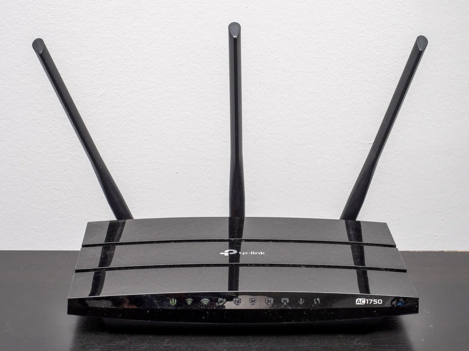 A black home Wi-Fi router with three upright antennas and a row of small front status lights, on a dark table against a white wall.